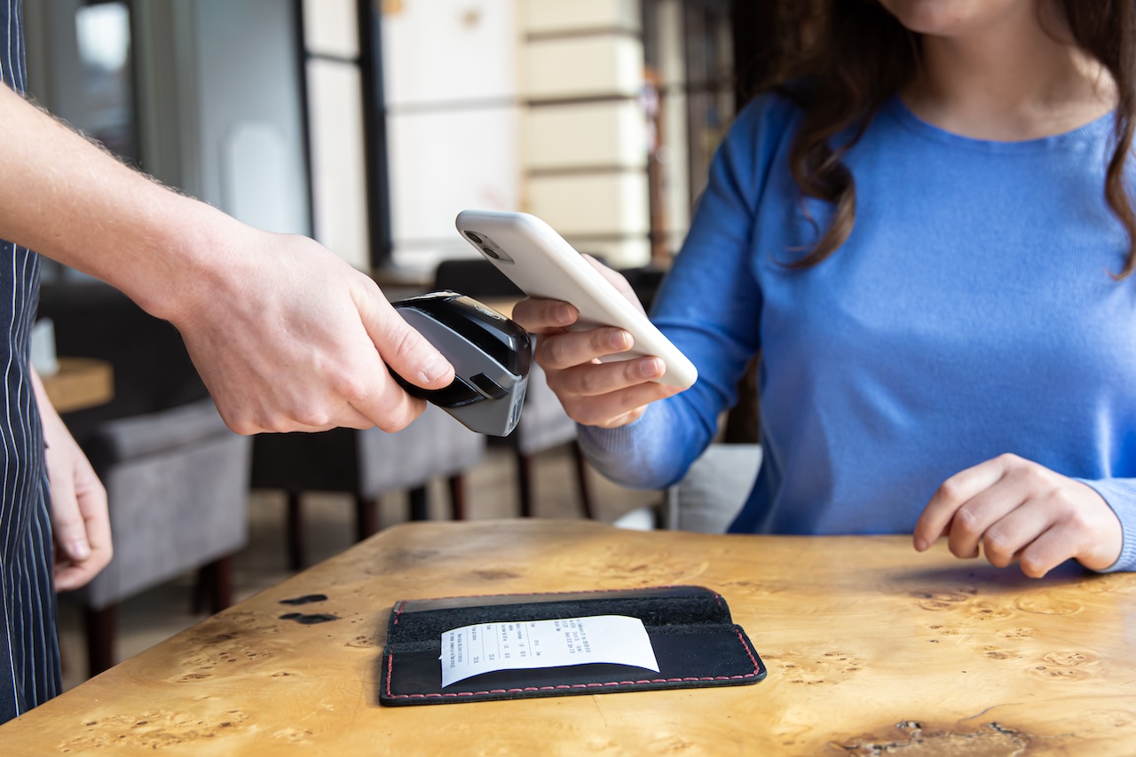 A woman sitting with a mobile smartphone and paying by pay pass, NFC technology on a cell phone and payment concept, indoor in cafe or restaurant.